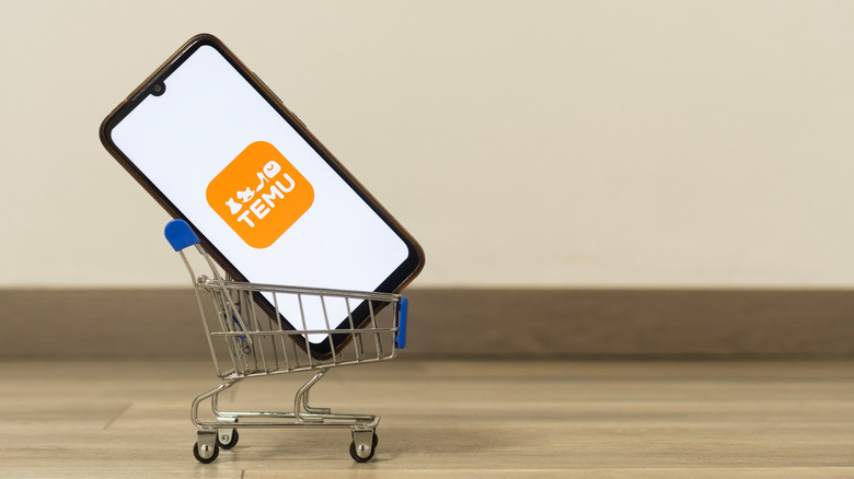 The temu logo on a smartphone placed on a miniature shopping cart