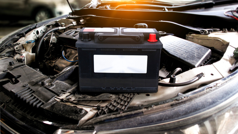 A black car battery sitting outside its housing with the engine hood open