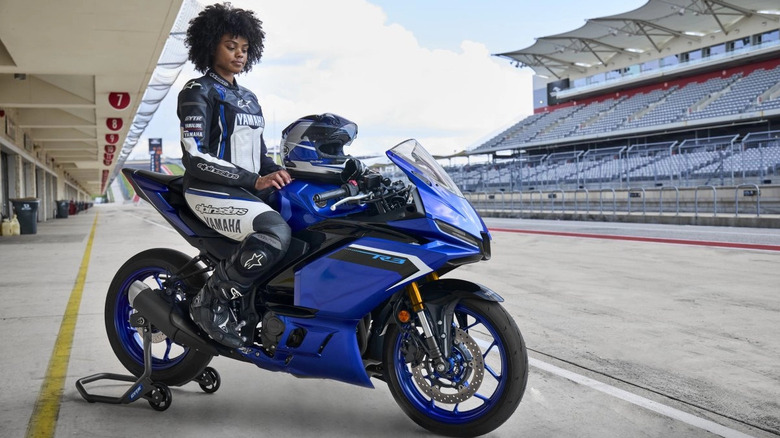 Female rider in full Yamaha racing gear sitting on a blue Yamaha YZF-R3 sportbike in a pit lane at a professional racetrack with empty grandstands in the background.