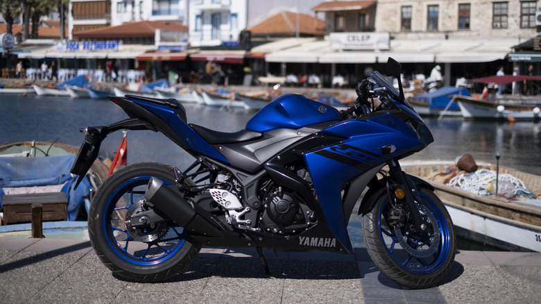 Blue Yamaha YZF-R125 sportbike parked by a harbor with fishing boats and waterfront buildings in the background on a sunny day.