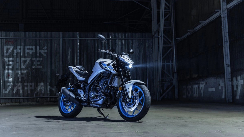 Yamaha MT-03 naked sportbike with blue rims parked in a dark industrial warehouse, illuminated by LED headlights with 'Dark Side of Japan' graffiti in the background.