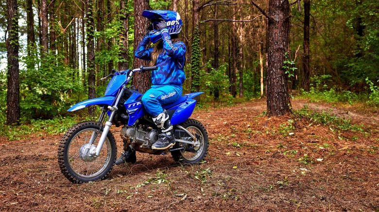 Young rider adjusting helmet while sitting on a Yamaha TT-R110E dirt bike in blue riding gear on a forest trail surrounded by tall trees and foliage.
