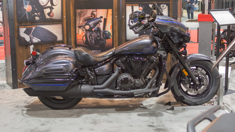 Yamaha V Star 1300 Deluxe touring cruiser motorcycle on display at an indoor expo, featuring hard saddlebags, batwing fairing, and custom accessories under showroom lighting.