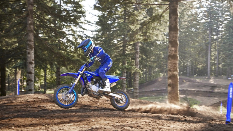 Young motocross rider in full Yamaha gear powers through a dirt track on a Yamaha YZ65, kicking up roost in a forested motocross park