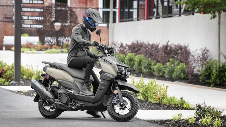 Rider in black gear sitting on a Yamaha Zuma 125 scooter in matte sand color, parked in a modern urban setting with landscaped greenery and directional signage in the background.