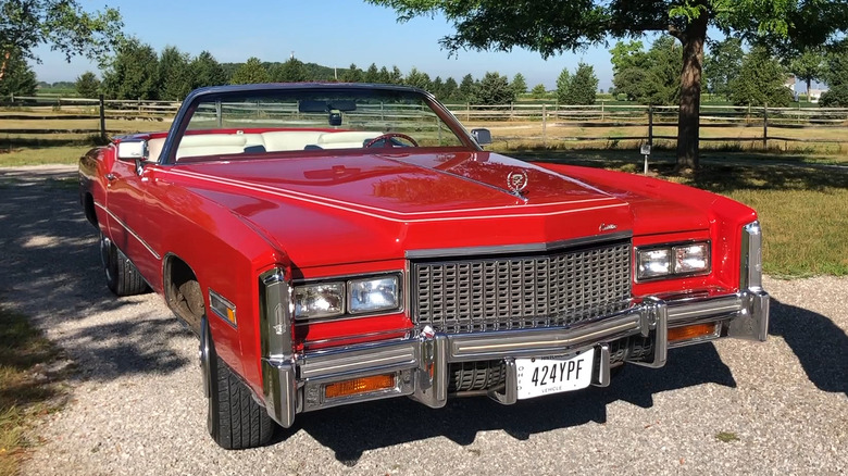 A 1976 Cadillac Eldorado convertible open-top cruiser finished in bright red, standing off a small country road in the shade.