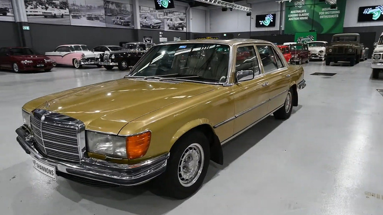 A green-gold 1970s Mercedes-Benz 450SEL 6.9-liter car parked inside a warehouse with other cars in the back.
