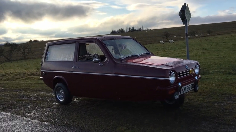 A red 1970s Reliant Robin parked off of the paved road on grass, with an agricultural field in the back.