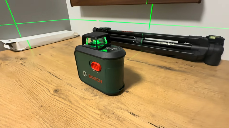 A green Bosch AdvancedLevel 360 Laser Level sitting on a desk while turned on, with a tripod visible in the back.