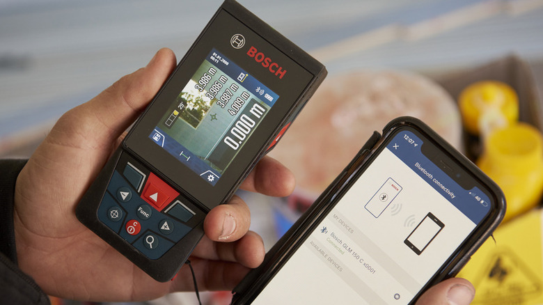 The Bosch GLM 150-27C laser measuring tool held in a man's hand next to an Apple iPhone with the Bosch app open.