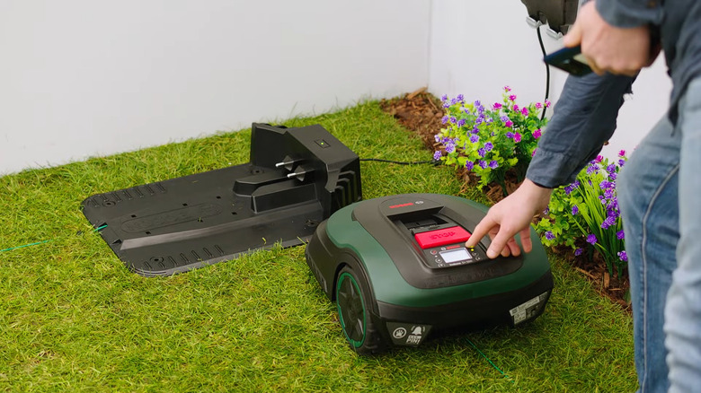 A dark matte green Bosch Indego LogiCut robotic lawn mower next to its docking station outdoors in a lawn