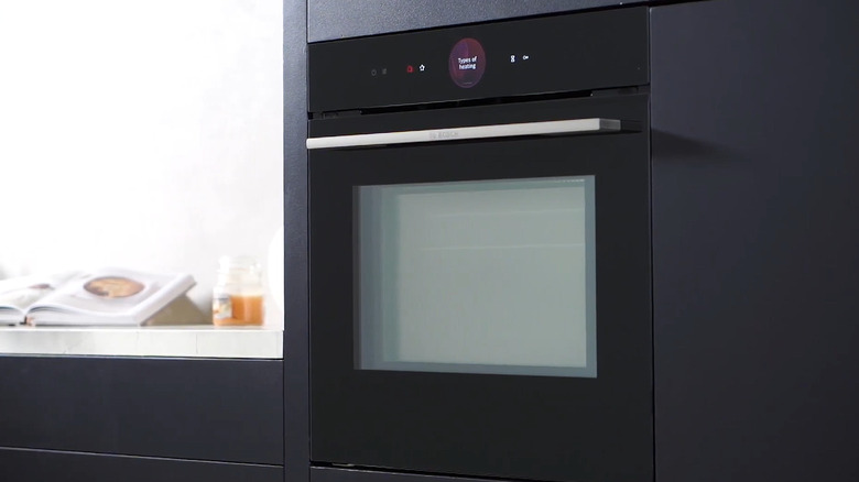A black Bosch Series 8 smart oven inset into the cabinet wall of a modern kitchen, also finished in a dark color like the oven.