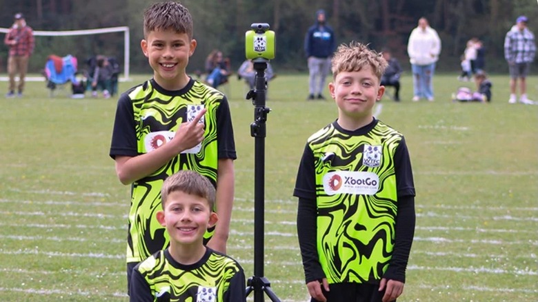 Three male children standing next to a camera on a tripod