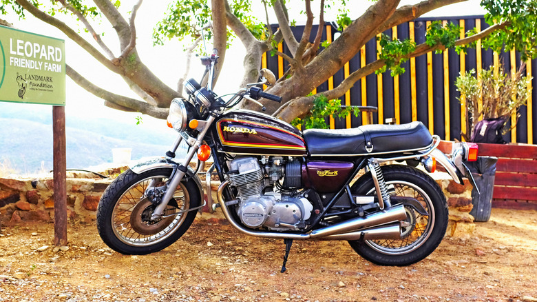 Classic Honda CB750 Four K motorcycle in a vivid maroon and gold finish, parked under a tree near a "Leopard Friendly Farm" sign.