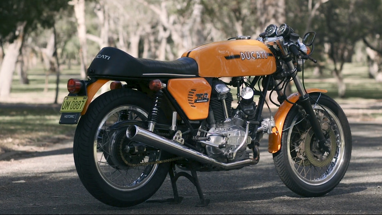 1974 Ducati 750 Super Sport in classic orange and black livery, elegantly displayed in a wooded park setting.