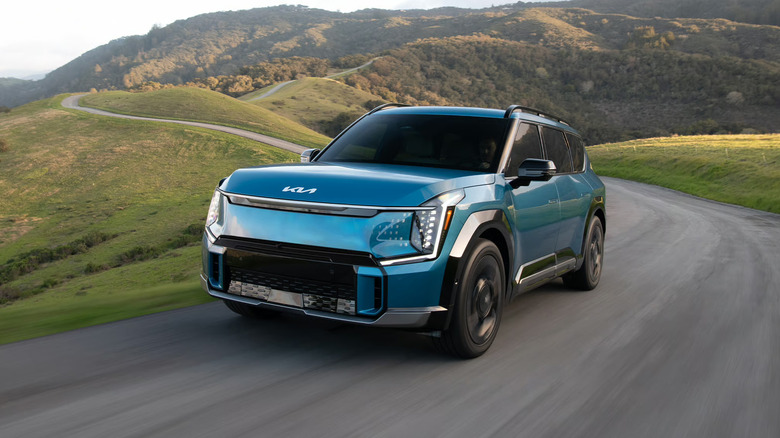 A light metallic blue Kia EV9 all-electric full-size SUV taking a corner on a rural road at speed, with hills and a forest in the backdrop.