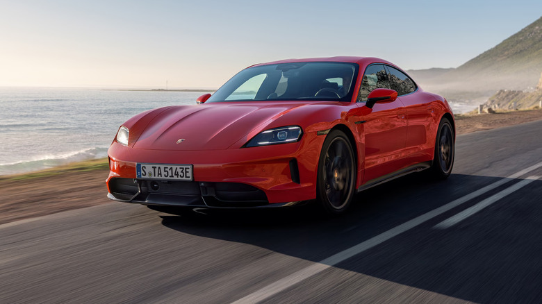 A metallic-finished red Porsche Taycan for the 2026 model year speeding down a coastal road at dusk.
