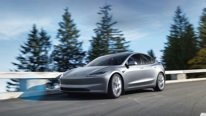 A silver Tesla Model 3 sedan taking a corner at speed in a green natural environment, left-side view visible.