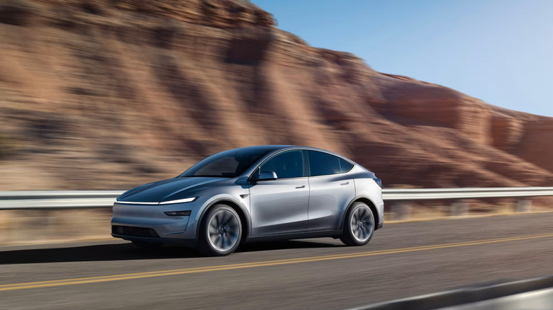 A silver Tesla Model Y for the 2026 model year traveling at speed down a highway, with red stone hills in the back.
