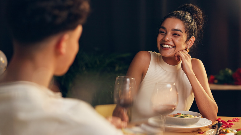 A woman sitting at a restaurant smiles to a person that is out of focus.