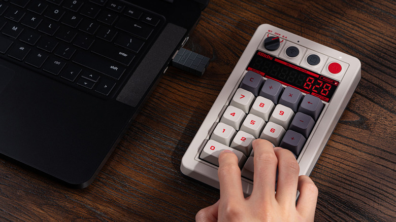 A person with their fingers on the 8BitDo Retro 18 Mechanical Numpad, placed on a wooden surface next to a laptop