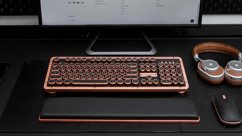 The Azio Retro Classic Bluetooth keyboard in front of a monitor, with a pair of headphones, a mouse, and a wrist wrest placed around it on a black surface