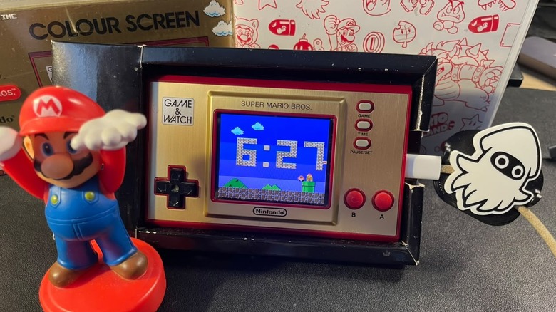 The Super Mario Bros. Game & Watch in its cardboard case, plugged into a charger with a Blooper pin next to a Mario figurine