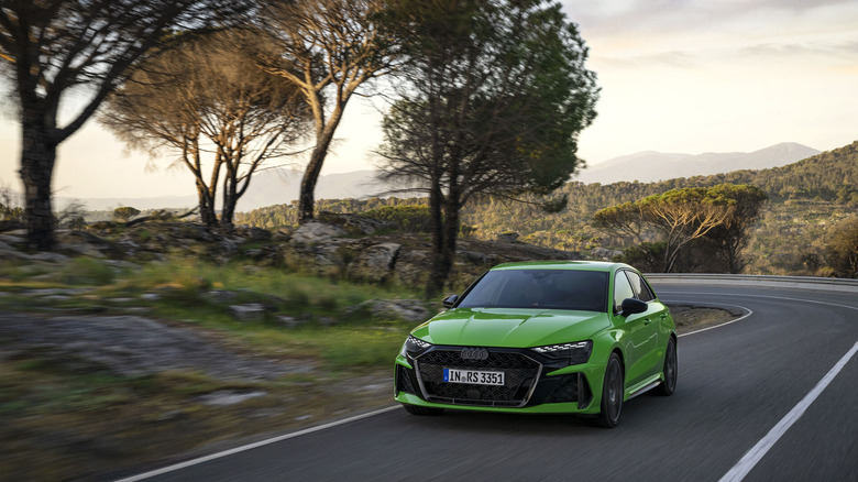 A bright green Audi RS3 Sportback hatchback car coming out of a turn on a curved road with shrubbery on both sides.