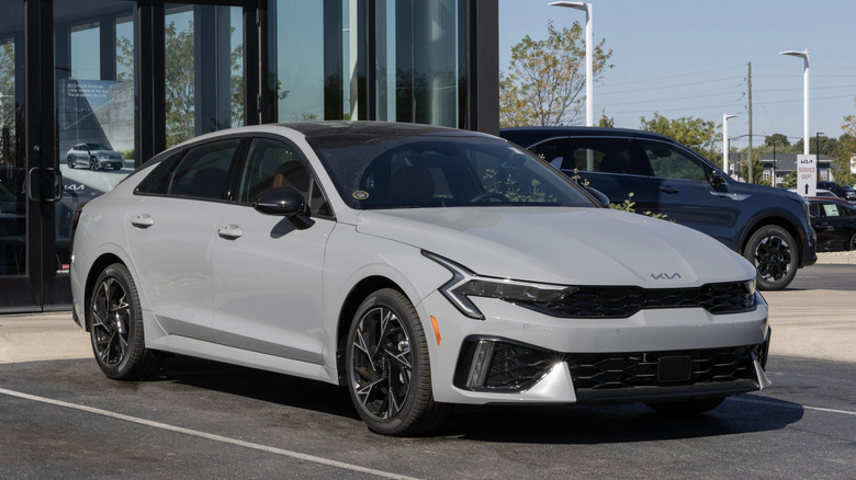 A gray 2025 Kia K5 GT-line on display outside a Kia dealership, with other cars in the background.