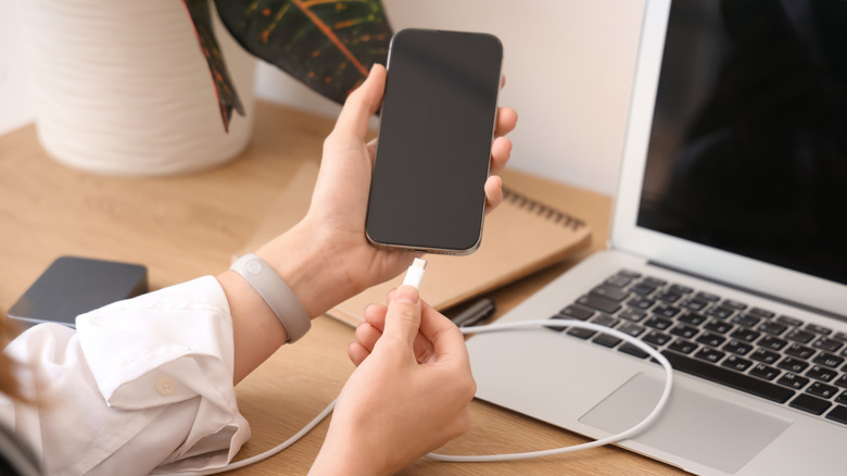 A person plugging their phone into charge with a laptop in the background