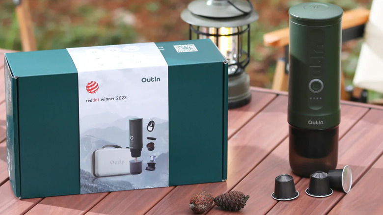 OutIn battery powered espresso machine on a table next to product box with camping lantern and chair in the background
