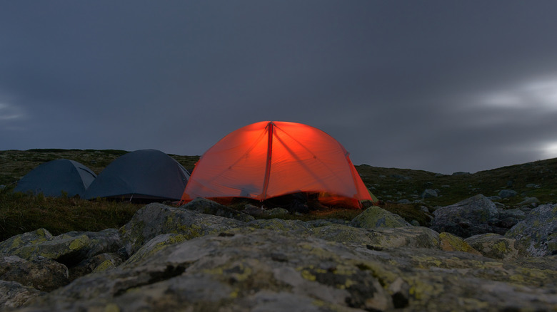 Tent with light glowing from within in the dark on rocky ground, with two unlit tents to the left of it