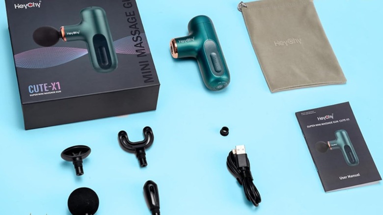 The Heychy Super Mini Massage Gun, disassembled with its body, heads, charging wire, case, and manual on a blue surface