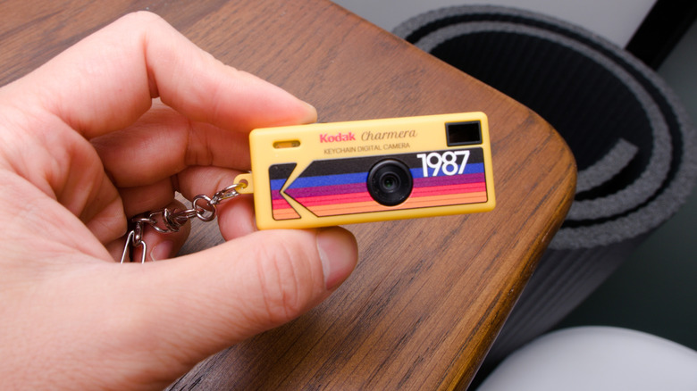 A hand holding the Kodak Charmera in front of a wooden desk, with a yoga mat next to it