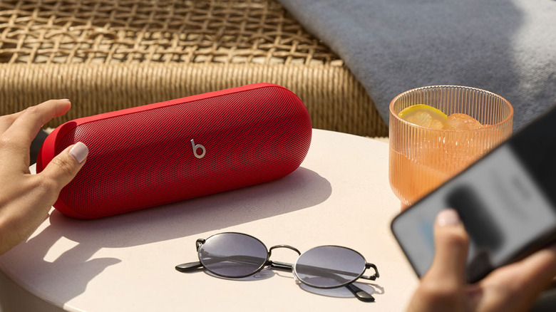A red Beats Pill placed on a table with shades and an orange drink, with a hand hovering over it while another holds a phone in the foreground