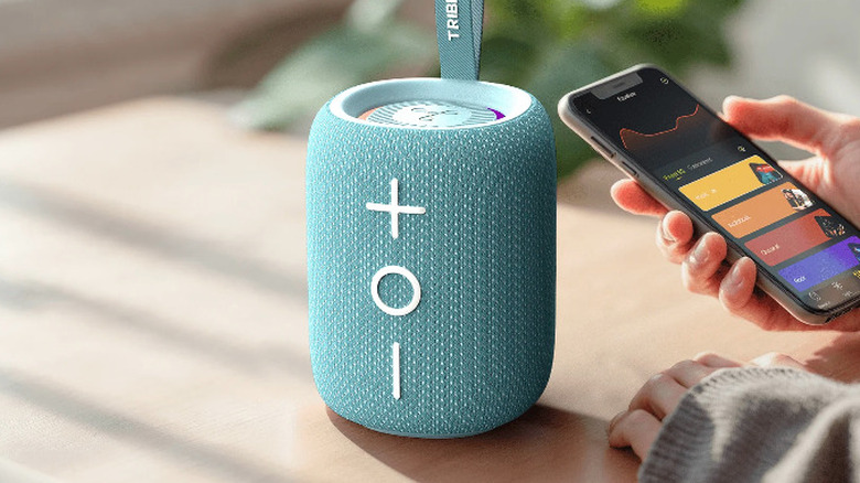 The Tribit StormBox Mini+ placed on a table, with a hand holding an app near it