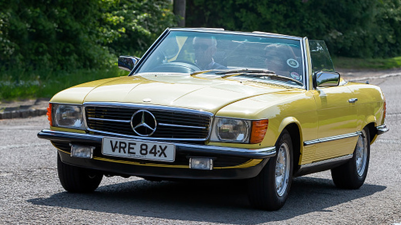 A classic yellow Mercedes-Benz SL-class roadster open top driving along through an English village.