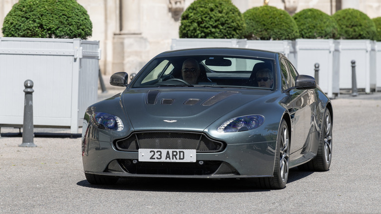 A silver-gray Aston Martin Vantage parked in front of a historic-looking, beige building in the UK.