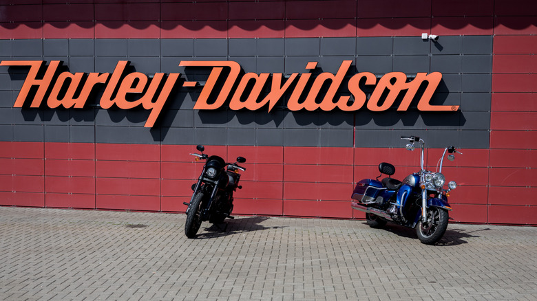 Two Harley-Davidson motorcycle cruisers parked in front of a large, orange Harley Davidson sign mounted on a red wall.