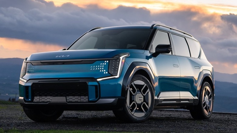 A blue Kia EV9 electric SUV parked on a hilltop at sunset.
