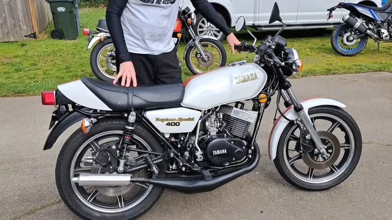 An RD400F Daytona Special sits outside a garage, surrounded by a few other bikes. Its owner, eager to take it for a spin, grips the bars, ready to bring the two-stroke legend to life.