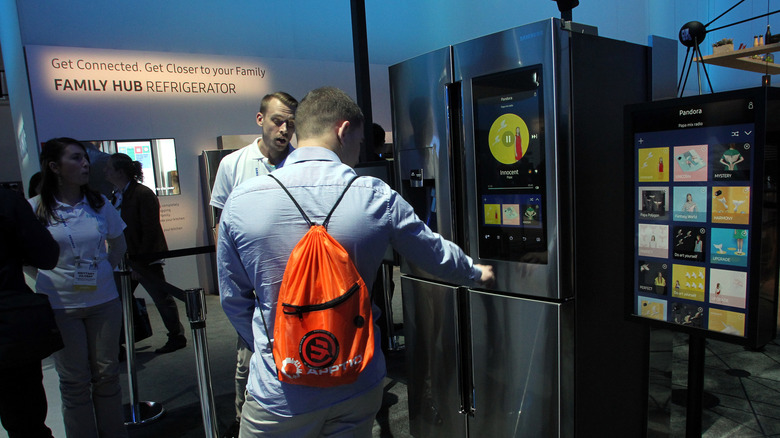 A silver-colored Samsung Family Hub fridge, with its 21.5 inch screen on, with people interacting with it.