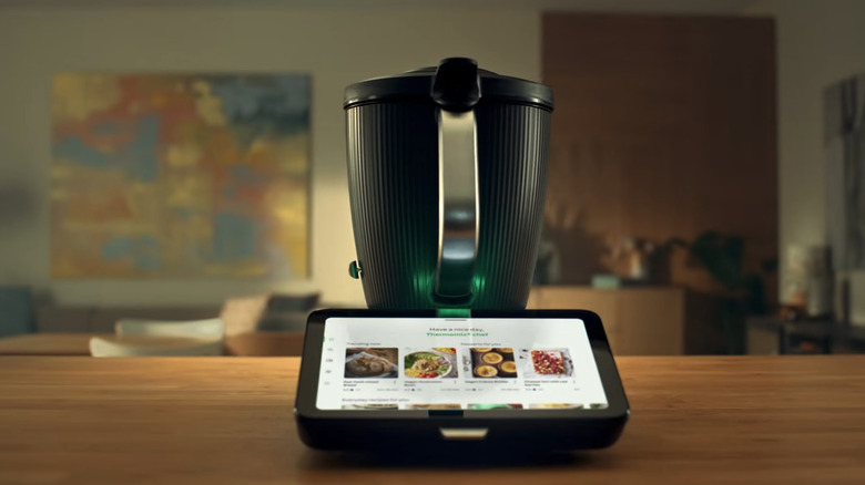 A black Thermomix TM7 cooking device in focus, sitting on a wooden countertop.