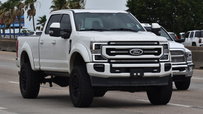 A white 2024 Ford F250 moving down a road in front of a RAM truck.