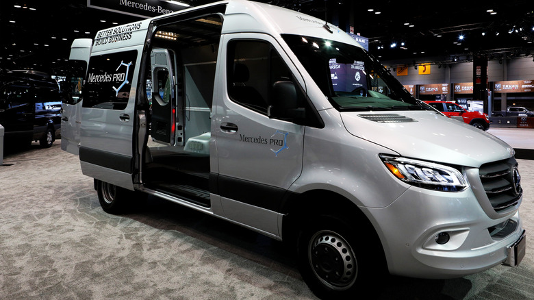 A silver Mercedes Sprinter passenger van with its sliding door open on display at an indoor motor show.