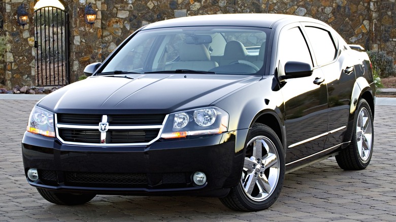 A 2013 black Dodge Avenger is parked on a stone-paved driveway in front of a rustic stone wall and gate.