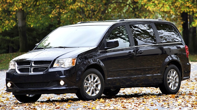 A 2008 black Dodge Grand Caravan is parked on a leaf-covered street surrounded by autumn trees.