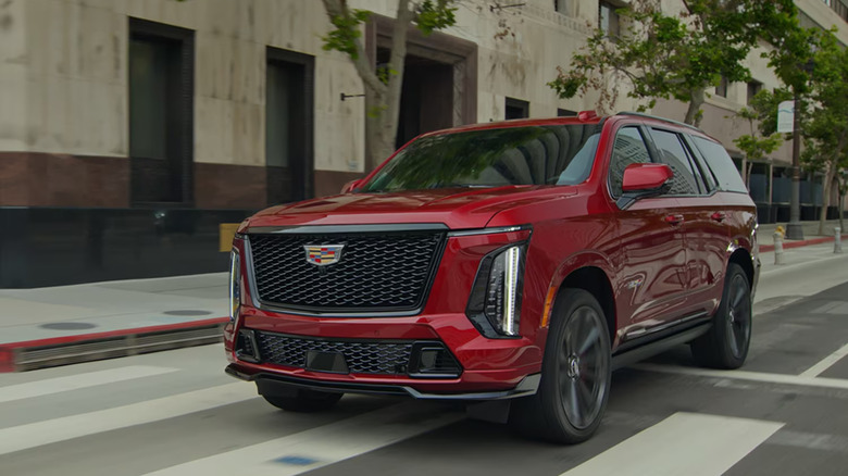 A red 2026 Cadillac Escalade V driving past a row of trees planted along an urban sidewalk