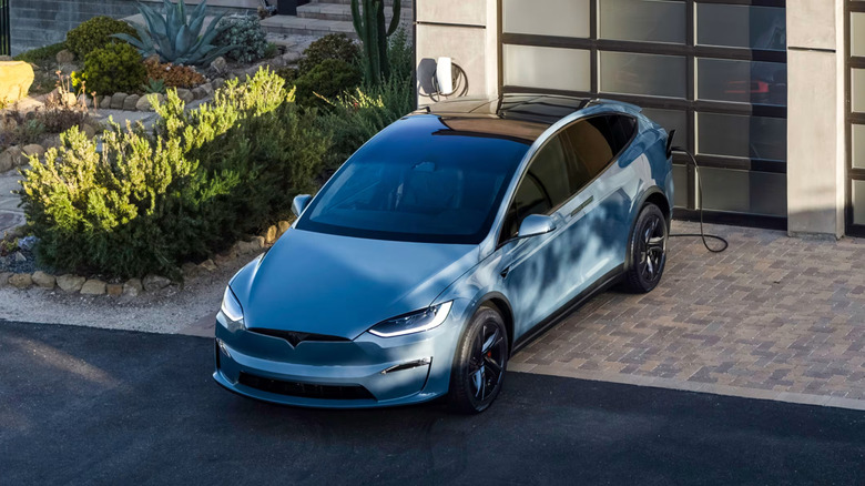 A light blue Tesla Model X charging in a driveway near a garage.