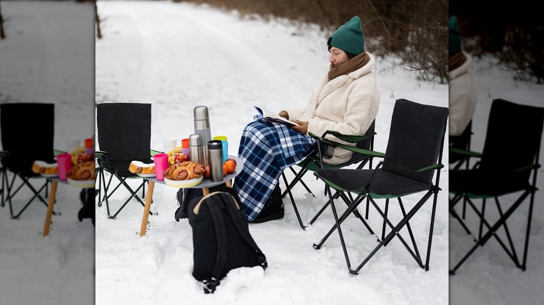 Woman reading on a black chair under a plaid blanket on a snowy backdrop